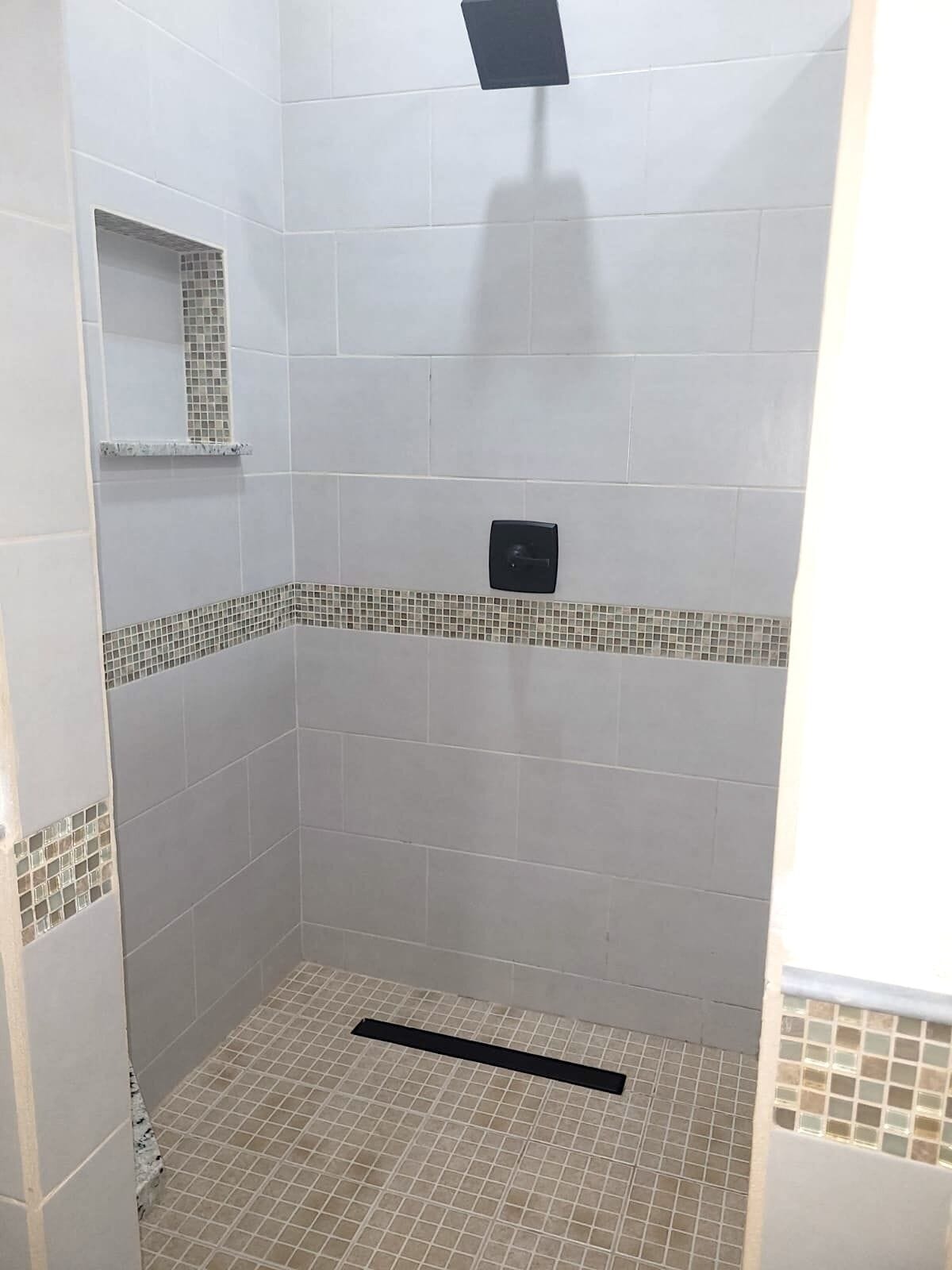 A small, enclosed shower area with tiled walls and floor, a shelf, and a control panel.