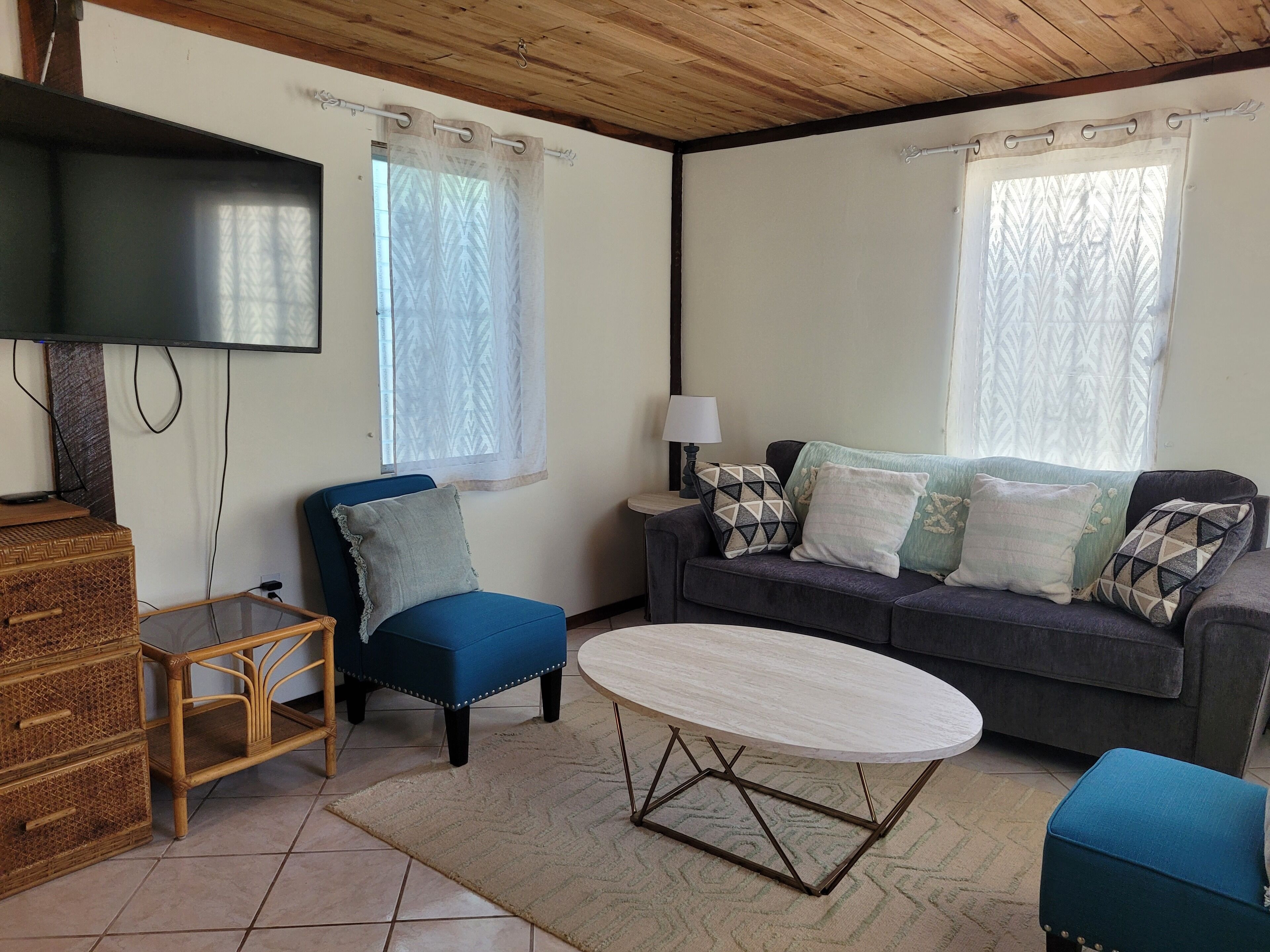 A living room with a blue armchair, a grey sofa with patterned cushions, a round coffee table, a television on the wall, and a wooden cabinet.