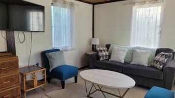 A living room with a blue armchair, a grey sofa with patterned cushions, a round coffee table, a television on the wall, and a wooden cabinet.