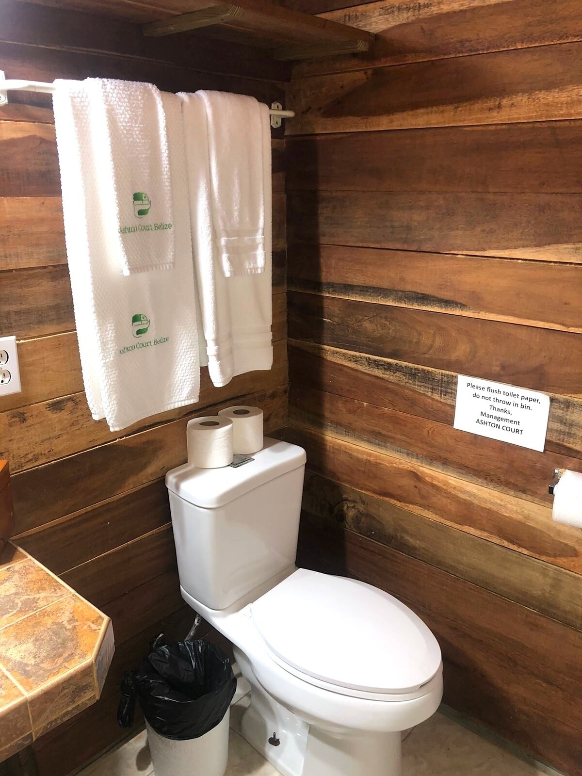 A wooden cabin-style bathroom with a toilet, two rolls of toilet paper, a black trash bin, and a sign instructing to flush toilet paper.