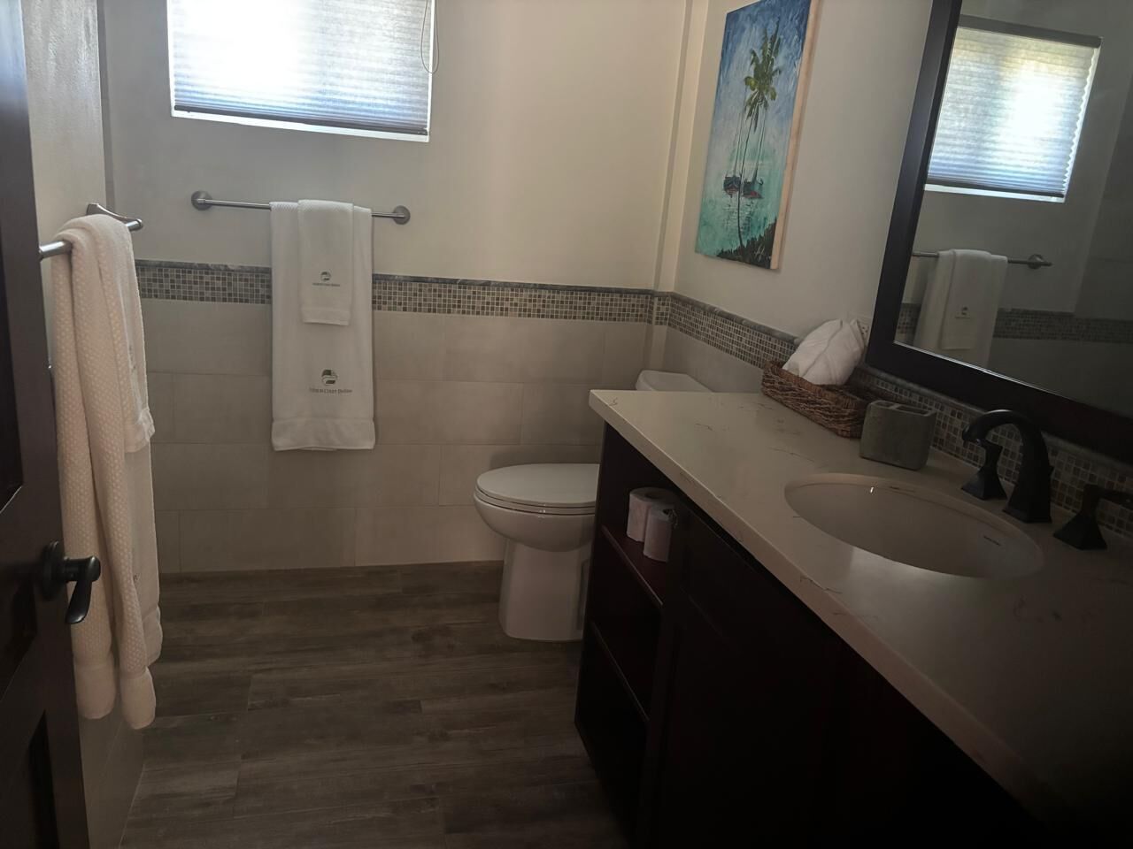 A bathroom with a toilet, sink, and mirror. There is a towel rack with towels, a window, and a painting on the wall.