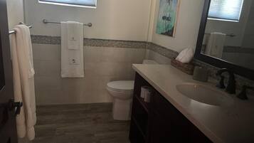 Un baño con inodoro, lavamanos y espejo. Hay un perchero con toallas, una ventana y un cuadro en la pared.