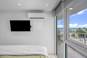 A bedroom with a bed, a mounted TV, an air conditioning unit, and a balcony with a view of the beach.