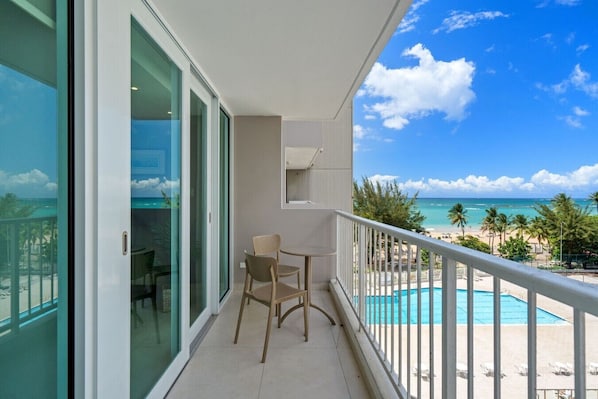 A balcony with a view of a beach, a pool, and a chair.