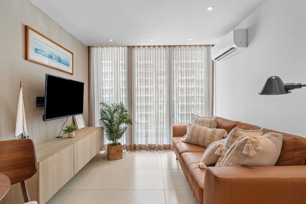 A modern living room with a flat-screen TV, a leather sofa, a potted plant, and a large window with sheer curtains.