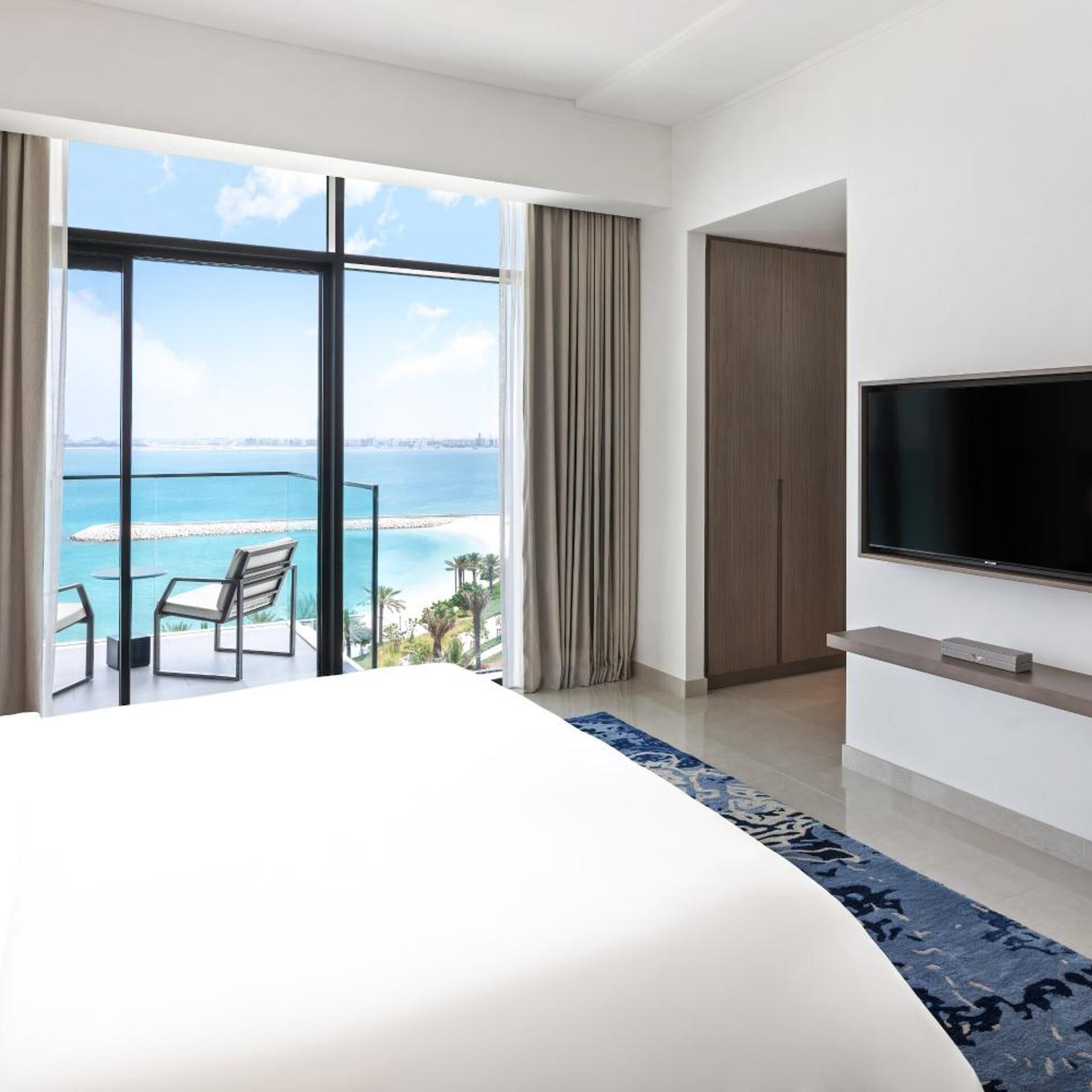 A hotel room with a large bed, a TV mounted on the wall, and a balcony with a view of the sea.