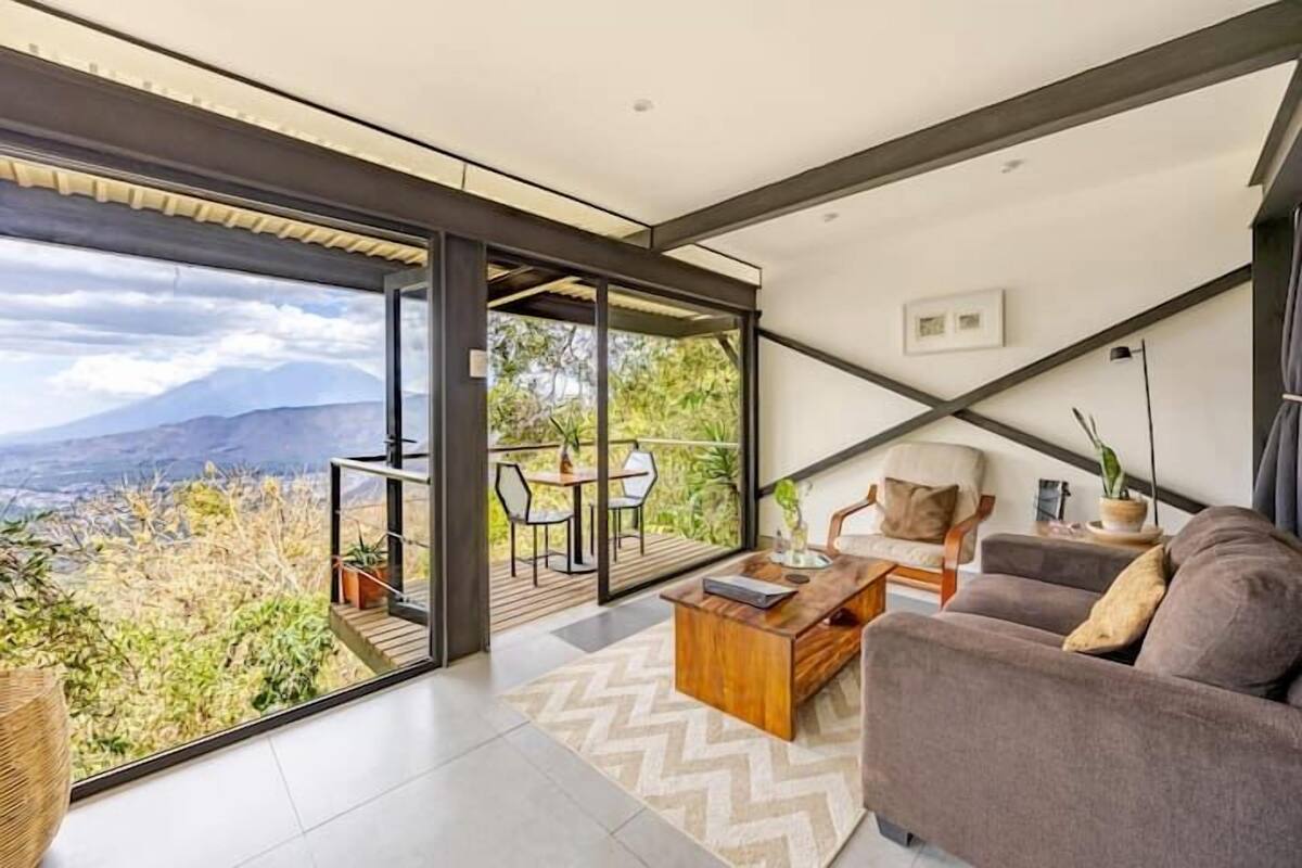 Een moderne woonkamer met een bank, een bijzettafeltje en uitzicht op een balkon met buitenmeubilair.