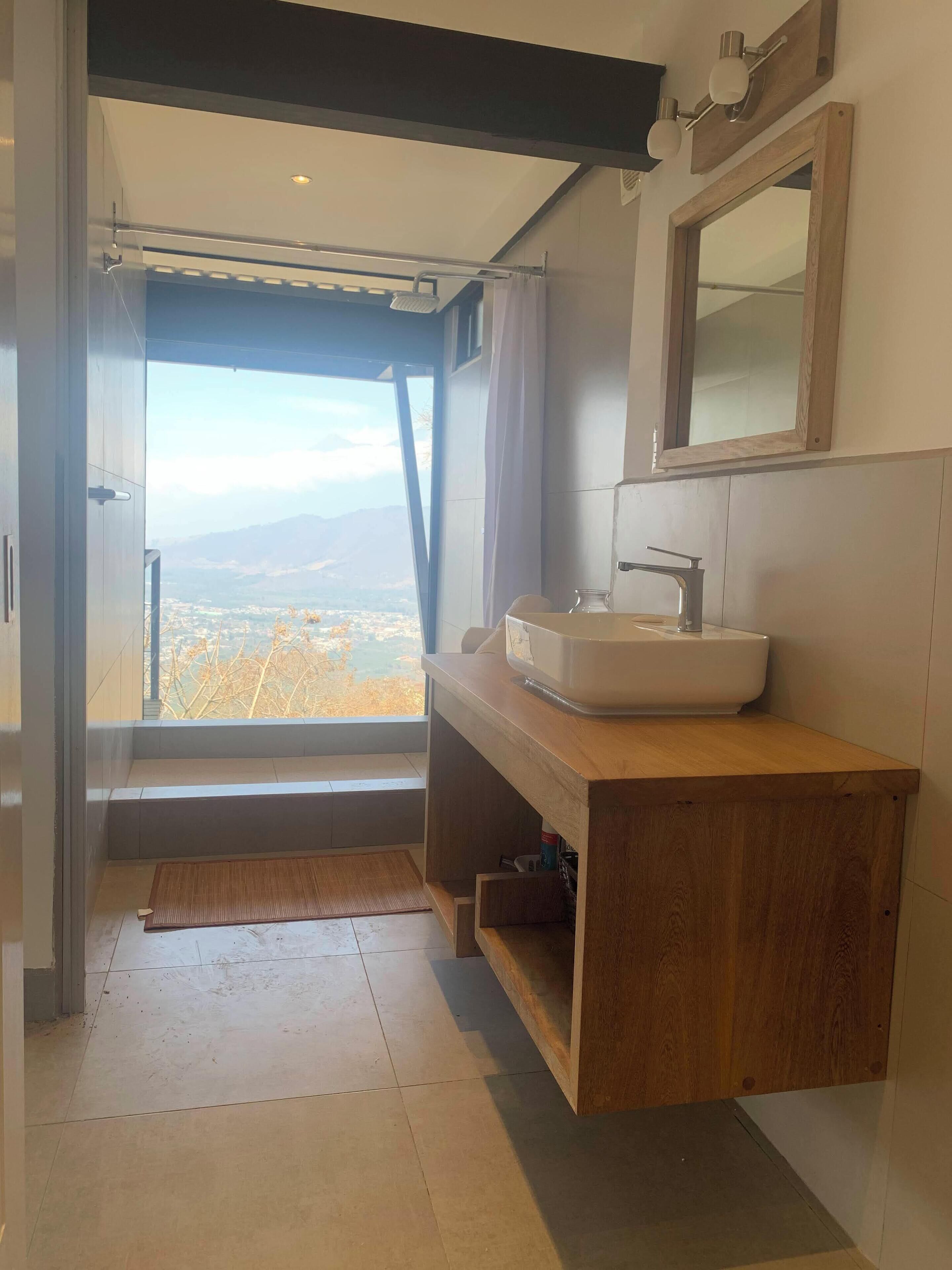 Een moderne badkamer met een houten wastafel, een grote spiegel en uitzicht op een bergachtig landschap door een open deur.