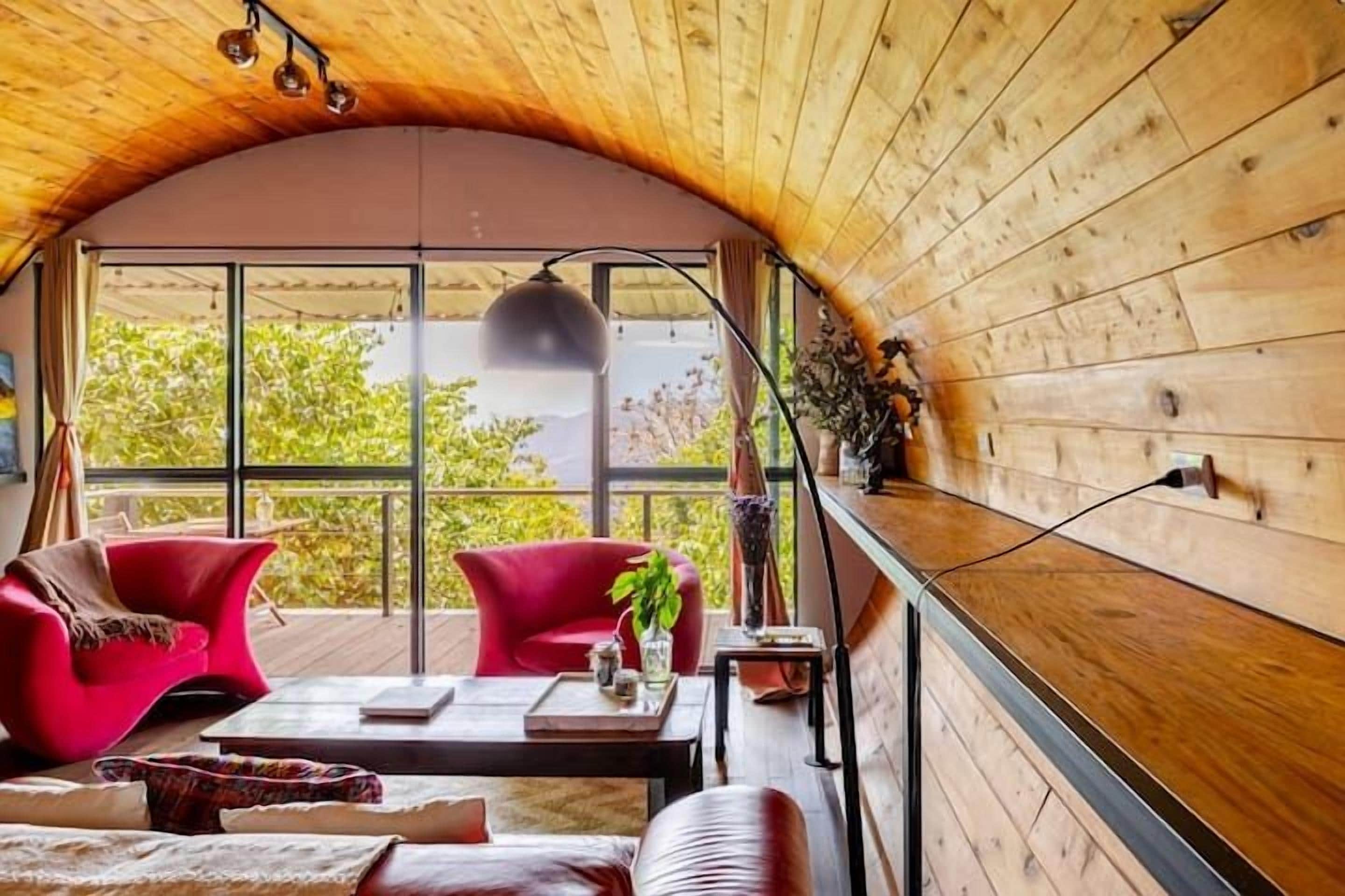 Una habitación acogedora con techo de madera curvada, ventanas amplias, sillas rojas, una mesa de centro y una planta.