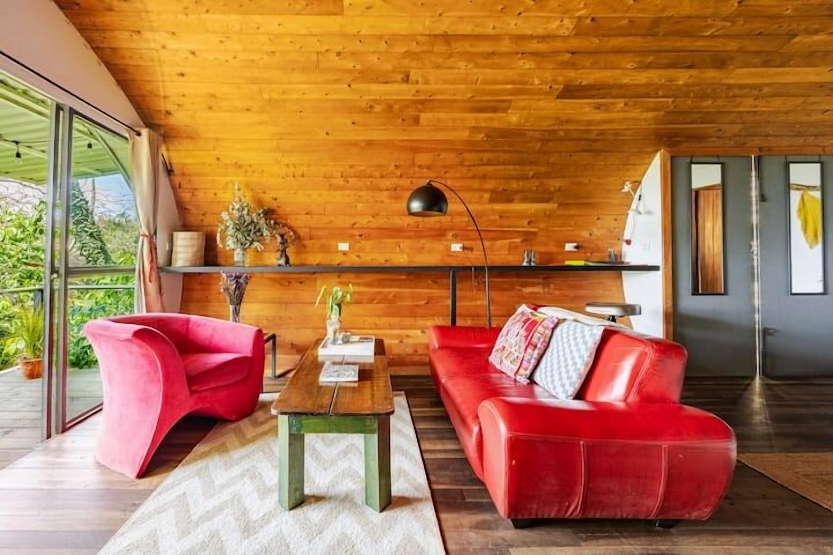 A modern living room with a wooden ceiling, a red sofa, a red chair, a wooden coffee table, and a floor lamp.