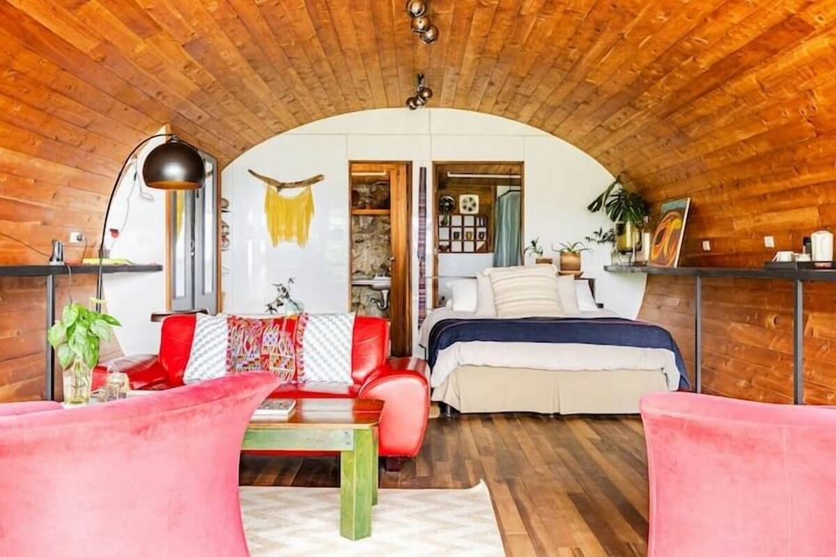 Een knus interieur met een houten plafond, een bed, een rode bank, een salontafel en een bureau met een lamp.
