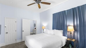 Ein Schlafzimmer mit einem Bett, zwei Nachttischen mit Lampen, einem Deckenventilator und einem großen Fenster mit Vorhängen.
