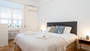 Uma cama bem arrumada com lençóis brancos, uma mesa de cabeceira com abajur e uma janela com persianas.