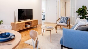 Uma sala de estar moderna com uma TV sobre um suporte de madeira, uma mesa de jantar redonda com pratos azuis, um sofá azul e uma cadeira com almofada floral.
