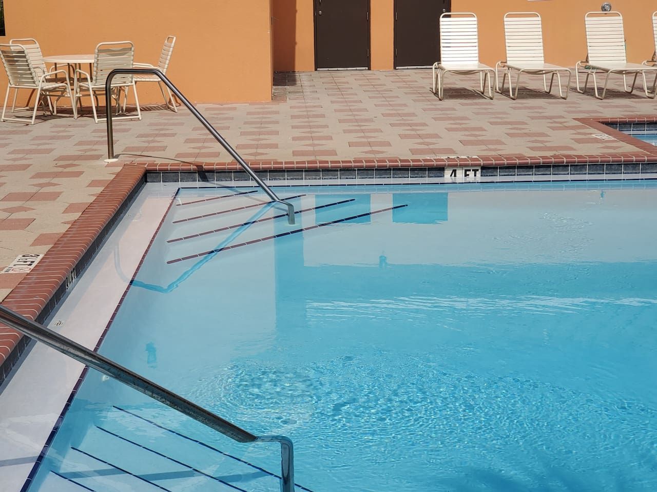 Una piscina con plataforma de baldosas, escalera de metal y sillas de descanso.