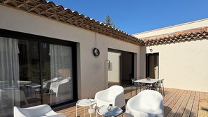 Une terrasse en bois avec des chaises et une table en plastique blanc, une porte-fenêtre coulissante et un toit recouvert de tuiles.