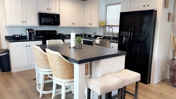 A modern kitchen with white cabinets, a black refrigerator, a microwave, and a coffee maker. There are two wicker chairs and a bench at the island. - Cozy 2BED 2BATH Home with central AC (Bacliff)
