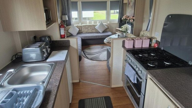 A compact kitchenette with a sink, stove, and a small seating area with cushions. - 3 Bedroom Caravan to Hire (Abergele)