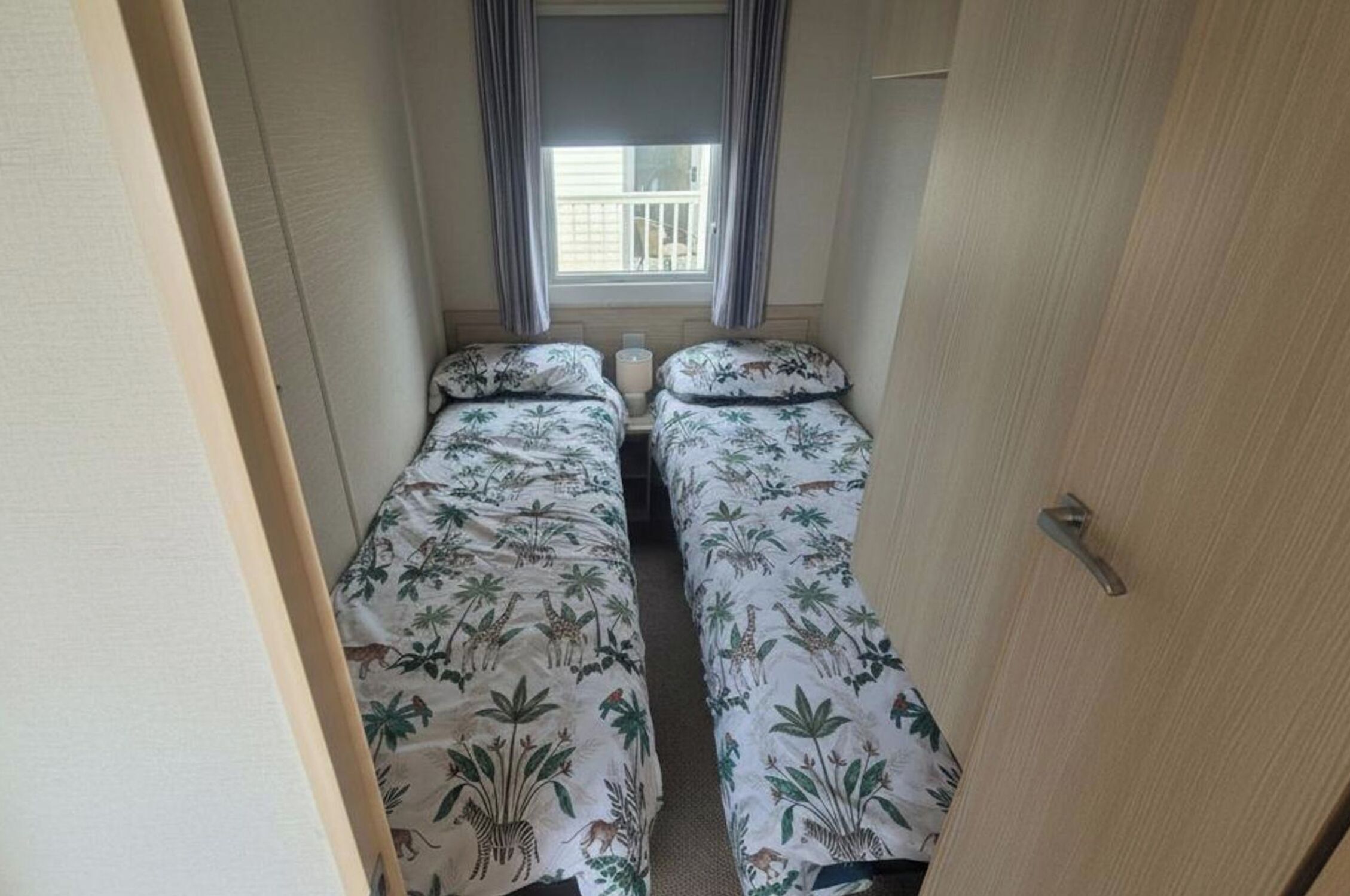 A narrow room with two beds, each with a floral bedspread, a window with curtains, and a door on the right.