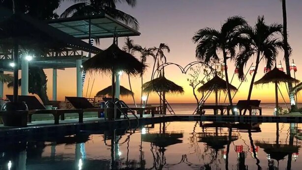 Un espace piscine paisible, doté de chaises longues et de parasols, où se reflètent le coucher de soleil et les palmiers.