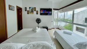 A hotel room with two beds, a ceiling fan, a TV mounted on the wall, and a large window offering a city view. - Estudios Mirador del Laguito (Cartagena)