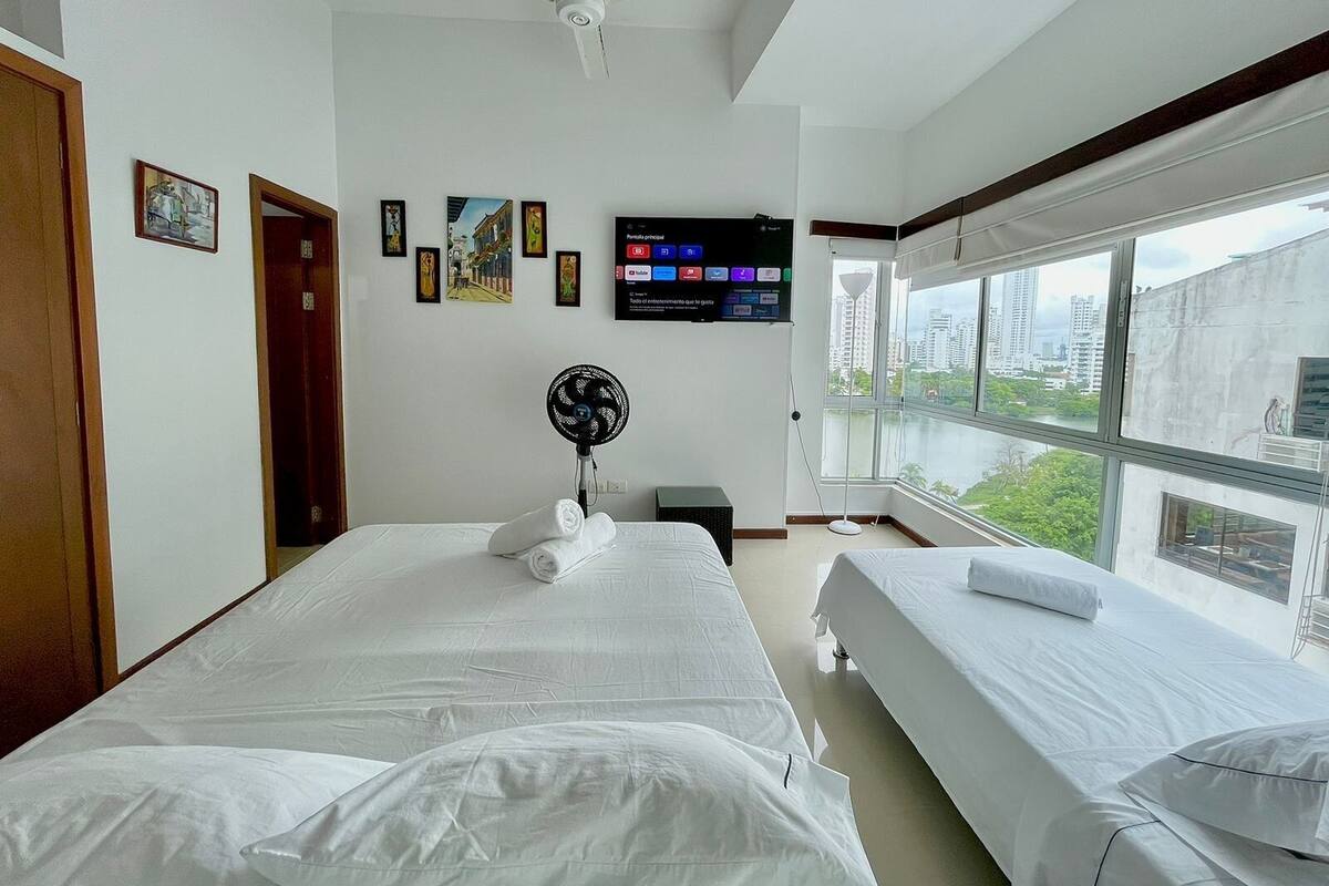 Habitación de hotel con dos camas, un ventilador de techo, un televisor montado en la pared y un amplio ventanal con vista a la ciudad.