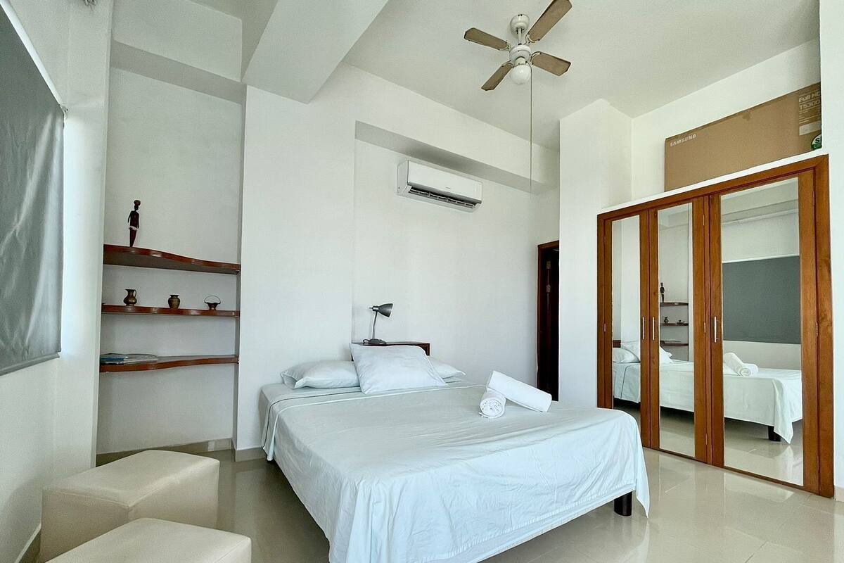 Habitación de hotel moderna con una cama grande, un ventilador de techo, un armario y un baño visible a través de una puerta de cristal.