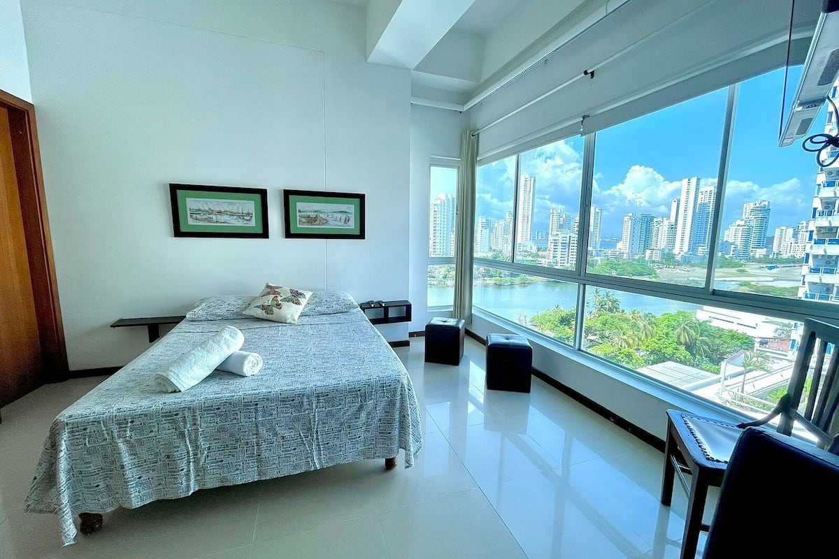 Un dormitorio moderno con una cama grande, dos cuadros enmarcados y vistas de un paisaje urbano a través de ventanas amplias. Se ve una vista de la ciudad desde el exterior.