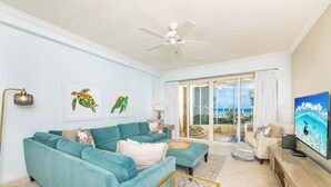 A living room with teal sofas, a ceiling fan, a TV, and a view of the ocean through large sliding doors.