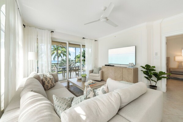 A spacious living room with a large sofa, a coffee table, and a television. There is a balcony with a view of palm trees and the ocean.