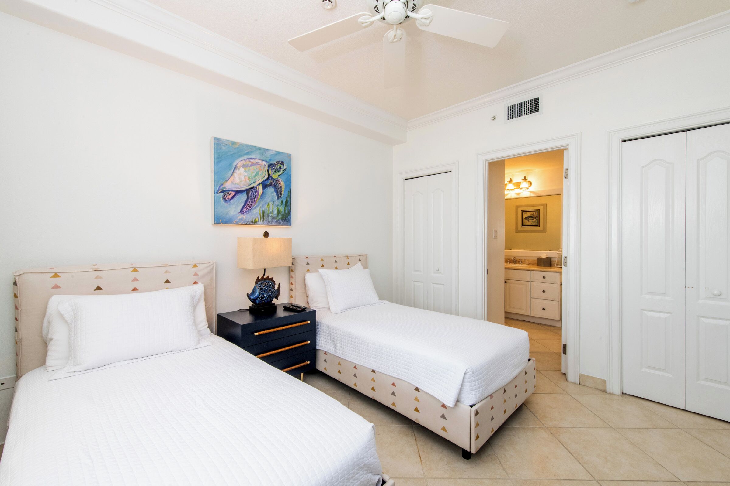A bedroom with two beds, a ceiling fan, a painting of a turtle, and a bathroom visible through an open door.