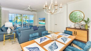 A spacious living room with a large window overlooking the ocean, a chandelier, and a dining area with a wooden table. - The Meridian, Villa 9, Second floor 3BR/3B, Oceanfront, fully equipped. (Seven Mile Beach)
