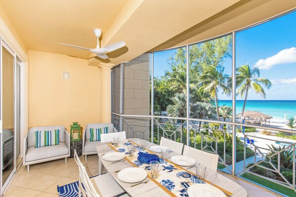 A dining area with a table set for four, a ceiling fan, and a view of the ocean and palm trees.