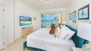 A bedroom with a bed, a ceiling fan, a TV, a window with a view of palm trees and the ocean, and framed artwork on the wall. - The Meridian, Villa 9, Second floor 3BR/3B, Oceanfront, fully equipped. (Seven Mile Beach)