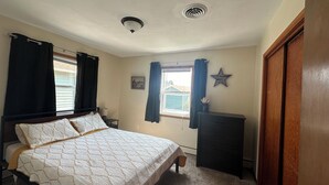 Ein Schlafzimmer mit Bett, Kommode, einem Fenster mit Vorhängen, einer Stern-Dekoration und einem Deckenventil.