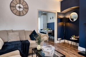 Ein modernes Wohnzimmer mit einem beigen Sofa, einem Couchtisch mit Büchern, einer Wanduhr und einem Essbereich im Hintergrund.