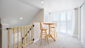 Una habitación con escalera de madera, una pequeña mesa con dos sillas y amplias ventanas con cortinas traslúcidas.