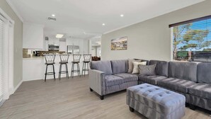 A modern living room with a grey sectional sofa, a matching ottoman, and a dining area with bar stools. - Spacious 4BR+Den w/ Pool, Jacuzzi, BBQ, Near Strip (Las Vegas)