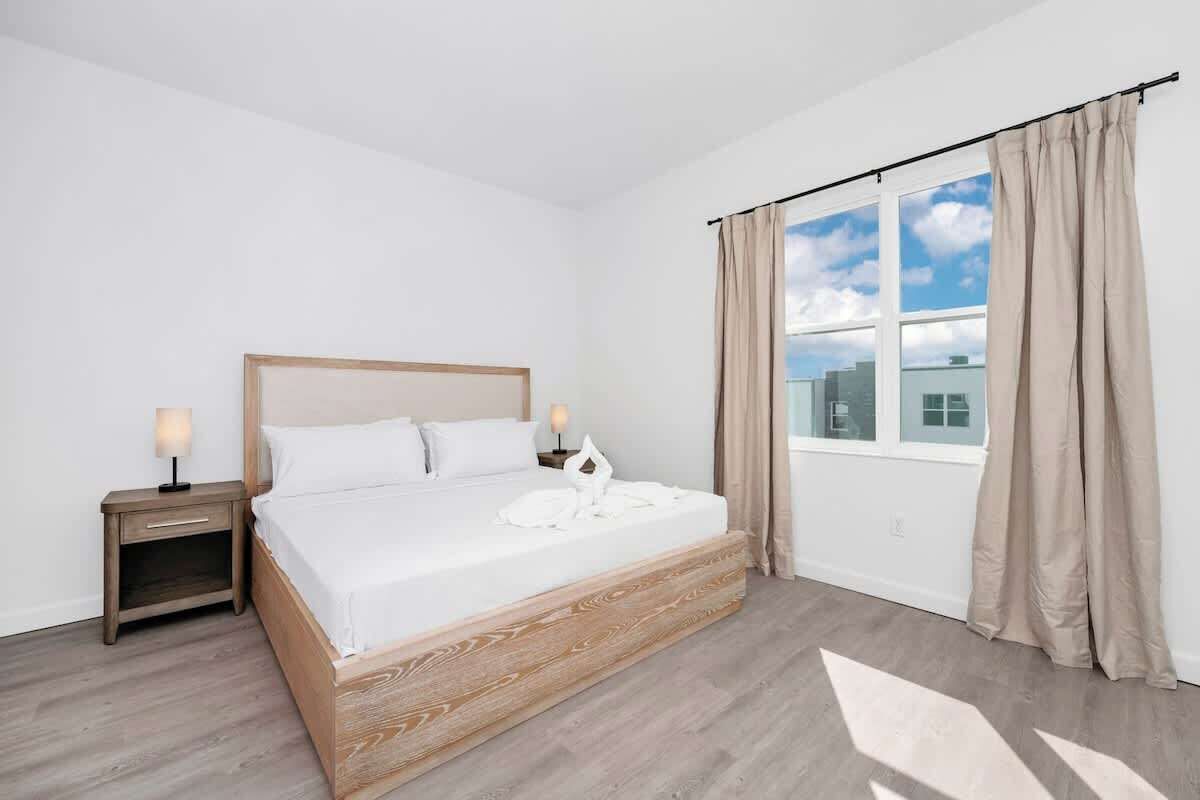 A modern hotel room with a wooden bed, a nightstand, a window with curtains, and a view of buildings outside.