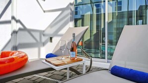 A sunlit outdoor seating area with a table, a bottle of wine, and glasses, near a large window with a view of buildings.
