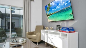 A modern living room with a large flat-screen TV displaying a beach scene, a beige sofa, a glass coffee table, and a white entertainment unit with board games.