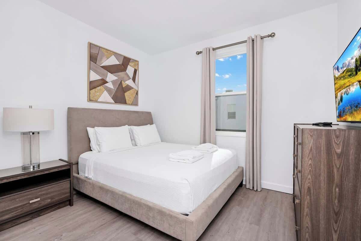 A modern hotel room with a large bed, a bedside table, a TV, and a window with curtains.