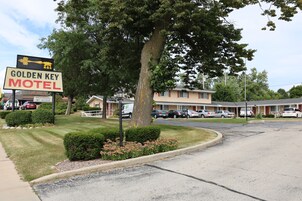 Un motel con un letrero que dice 'Golden Key Motel' y un letrero de 'Habitación disponible'. Varias automóviles estacionados adelante, y un edificio de techo rojo y paredes color beige.
