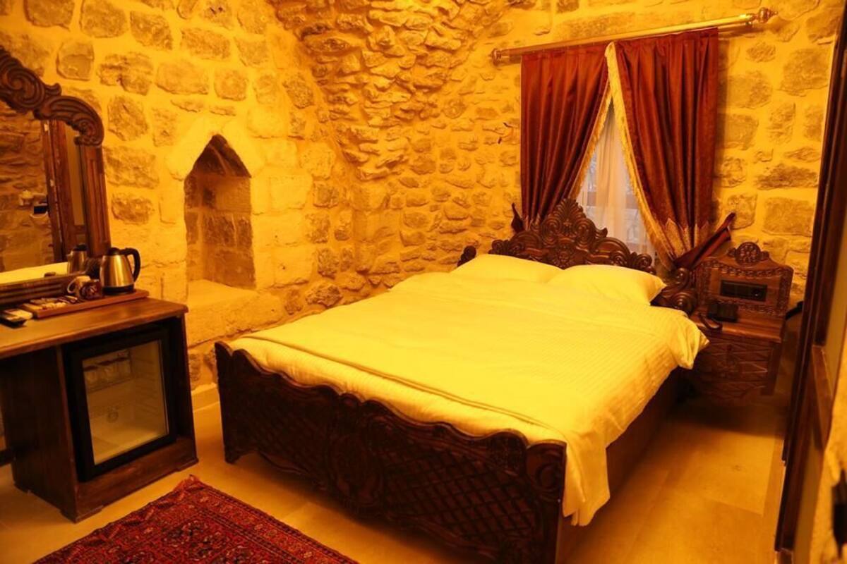Ein Schlafzimmer mit Stehwand, einem Holzbett, einem Nachttisch, einem Spiegel und einem Fenster mit roten Vorhängen.