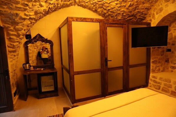 Un dormitorio con paredes de piedra, un armario de madera, una cama, un televisor y una pequeña mesa con una radio y una tetera.