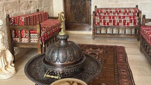 A traditional room with a stone wall, a wooden door, two wooden chairs with red and white patterned cushions, a large ornate metal tray with a cup of tea, and a rug with geometric patterns. - Kasim Efendi Konagi (Mardin)