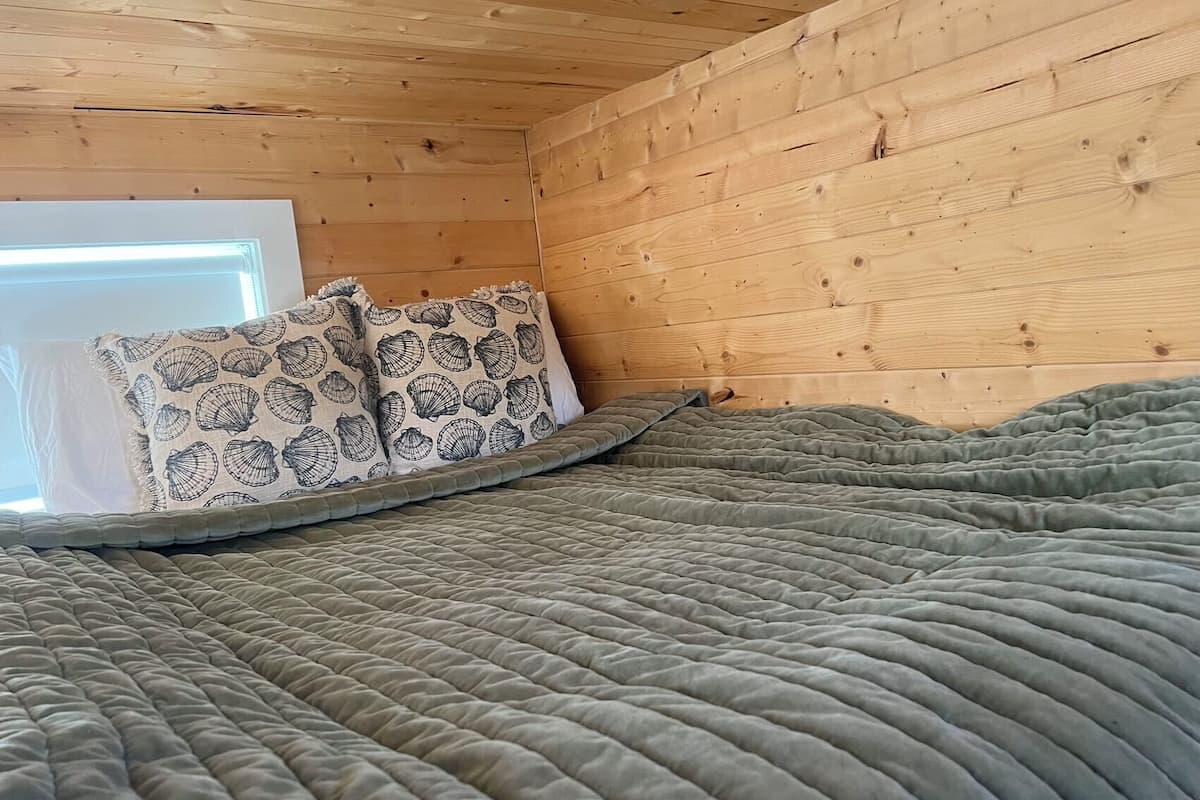 A cozy bedroom with a wooden ceiling, a bed with a grey quilt, and patterned pillows.