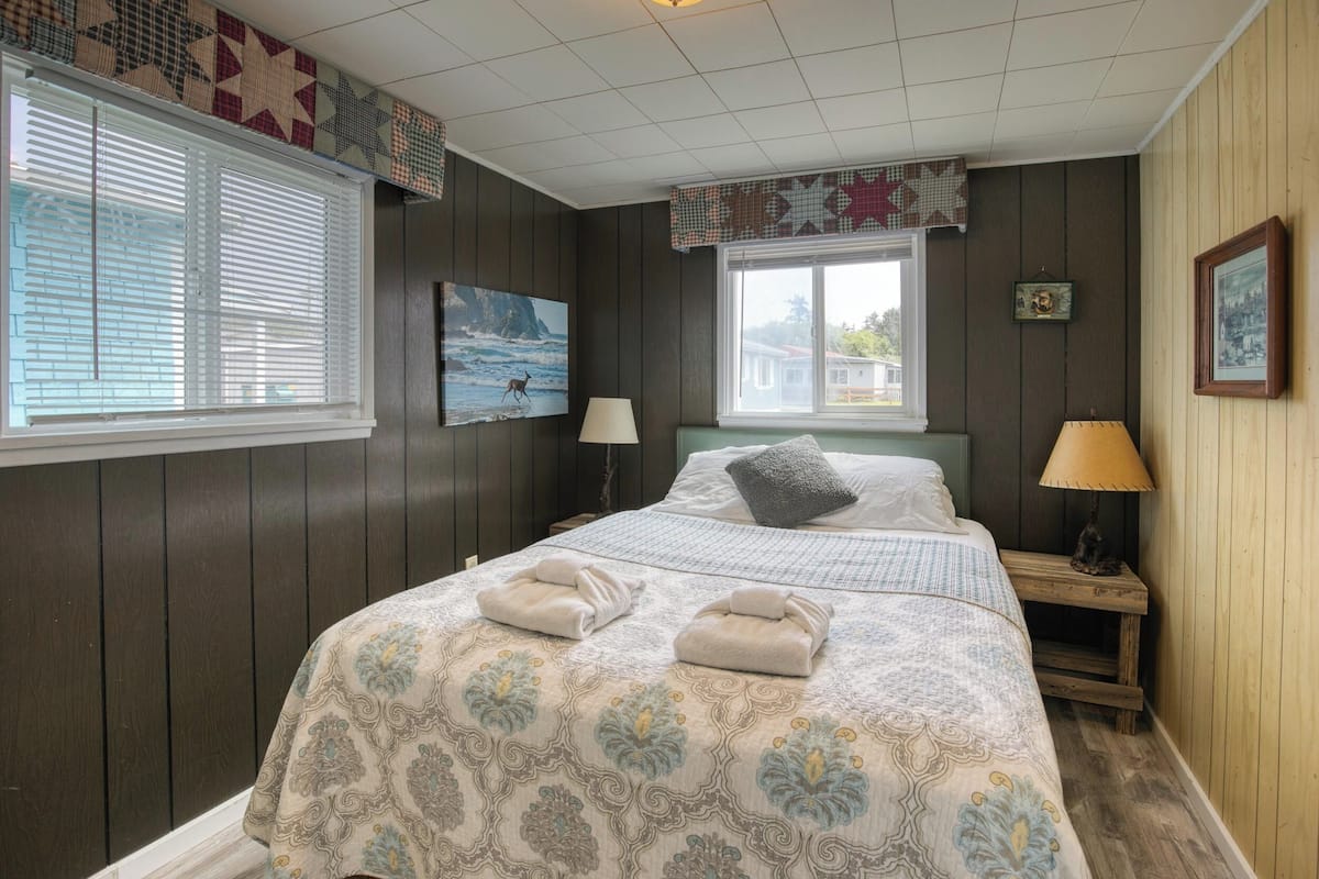 A bedroom with a bed, two bedside tables, a window with blinds, a lamp, and wall decorations.