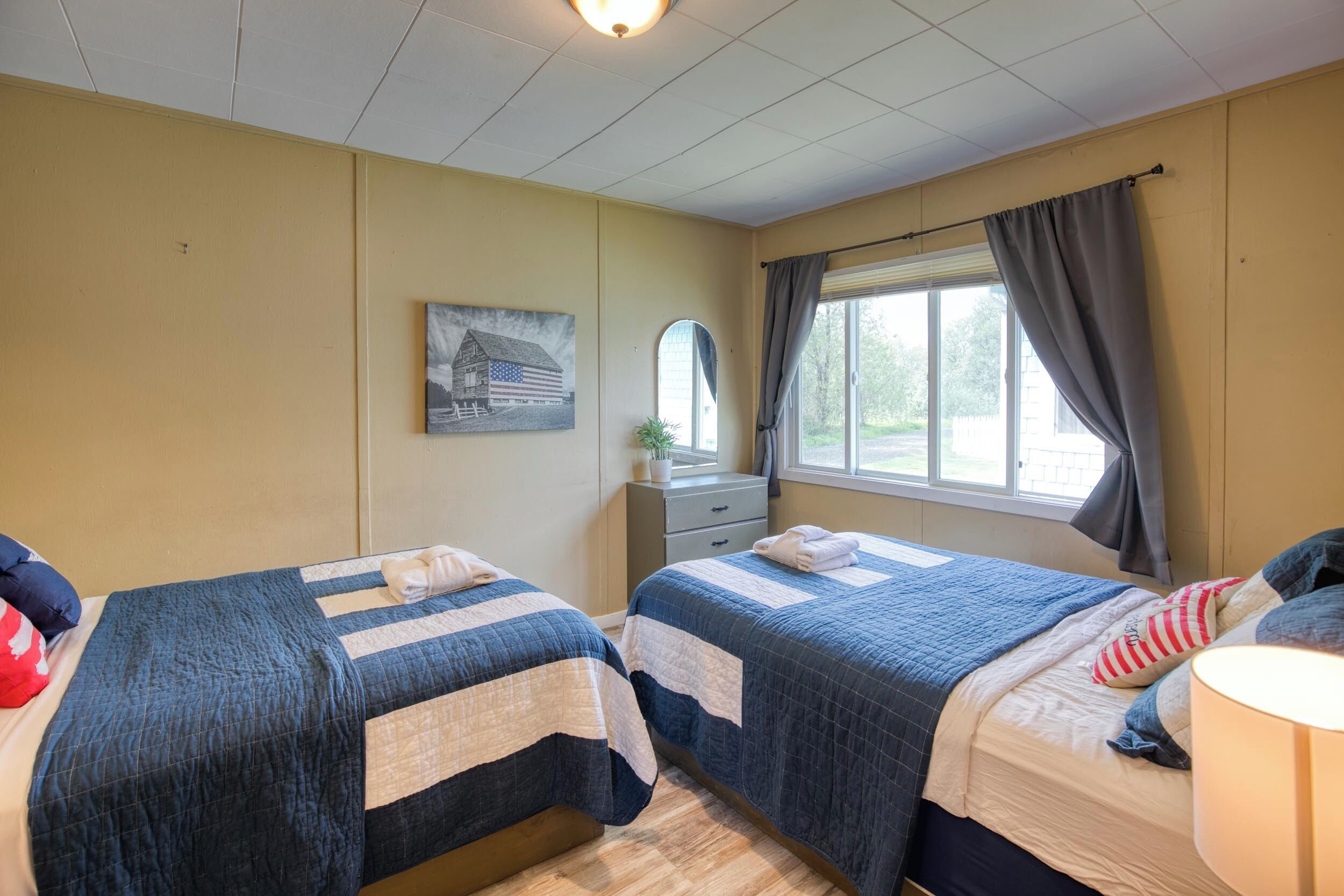 A hotel room with two beds, a window with curtains, a small desk with a mirror, and a wall-mounted picture.