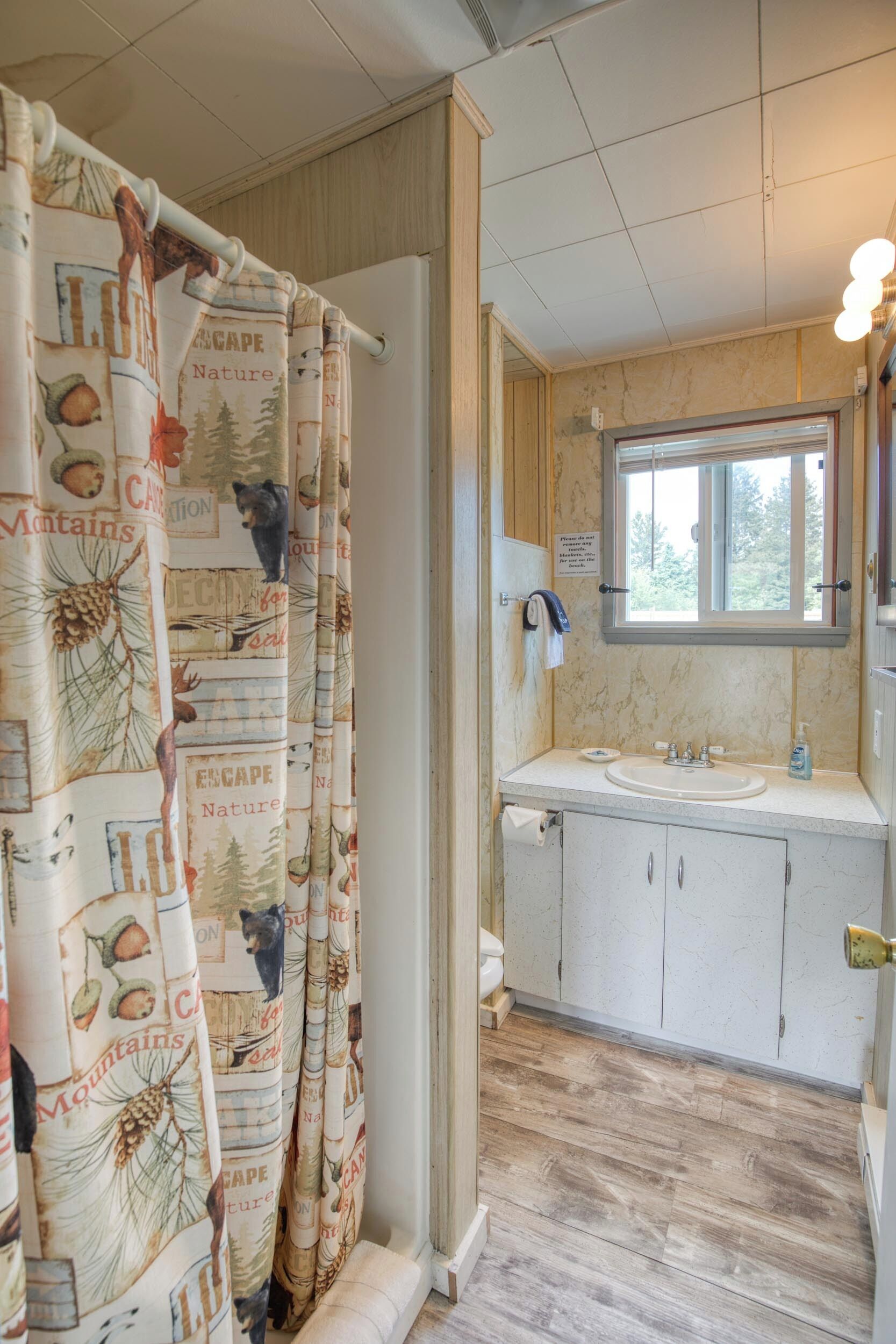 Un baño con cortina de ducha estampada con motivos naturales, una ventana sobre el lavabo y suelo de madera.